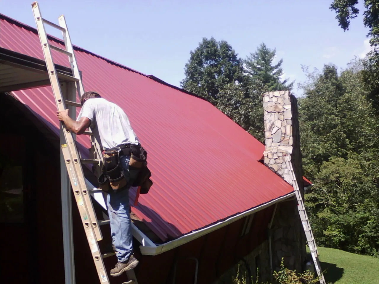 Roofer on a ladder beside a red metal roof and stone chimney — Soffit & Fascia Repair by Roofing Brothers in Coloma